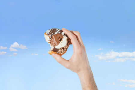 Summer creative scene with man's hand holding chocolate cake against pastel blue sky background with fluffy clouds. Concept of party catering, sugar addiction or eating sweet food.の写真素材