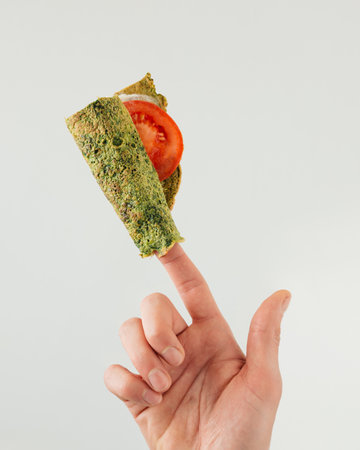 Man holds on finger rolled spinach omelette, tortilla or pancake, isolated on pastel grey background. Minimal concept of food, cuisine, ketogenic diet, low carb nutrition.の写真素材