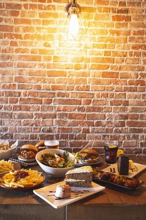 Variety of restaurant menu dishes. Salad with goat cheese, homemade hamburgers and fries, drink, chicken wings, vegetable tempura and cake on the wooden table. Vertical Imageの写真素材