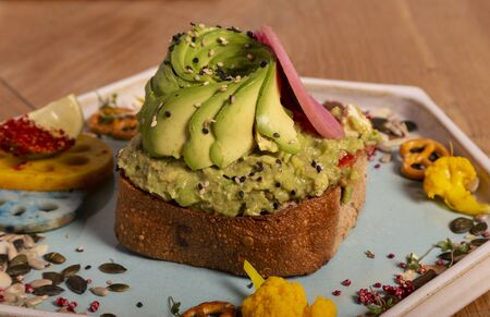 Close-up of toast with half avocado on a base of scrambled feta and avocado, grated tomato and oil served in an octagonal plate on wooden table. Artisan cookingの写真素材