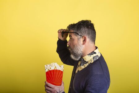 Portrait of attractive man with beard and sunglasses holding a box of popcorn in profile in amazed camera looking up at the glasses on yellow background.の写真素材
