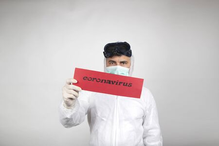 Man with surgical mask, glasses plus white protective suit and red sign with the word coronavirus isolated in studio on white background. New pandemic epidemic new fast-spreading coronavirus 2019-ncov. Medicinal flu virus concept.の写真素材