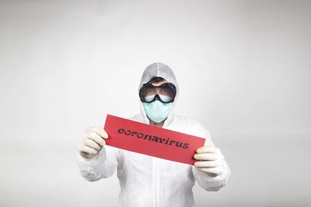 Man with surgical mask, glasses, white protective suit and red sign with the word coronavirus isolated in studio on white background. New pandemic epidemic new fast-spreading coronavirus 2019-ncov. Medicinal flu virus concept.の写真素材