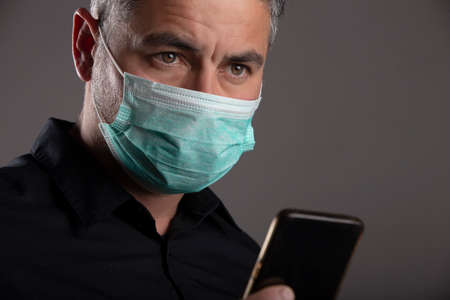Man with surgical protection mask with worried face after watching news on mobile on gray...
