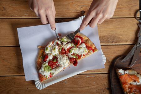 Woman's hands cut a piece of pizza on a wooden table. Italian pizza is eaten with a fork and knife....