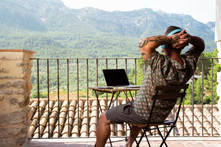 Handsome man relaxing and working on a laptop on the balcony of his house, while listening to music and enjoying the mountain viewsの写真素材