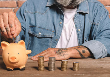 Man putting coins into piggy bank with stack of coins on table to save money and financial conceptの写真素材