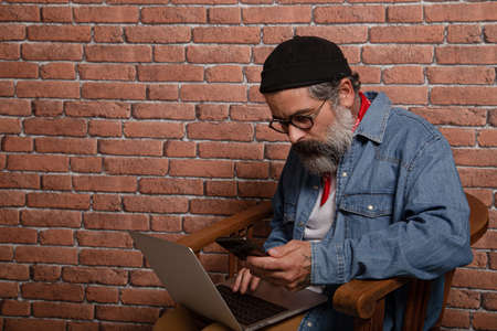 Side view portrait of cheerful bearded brunette man in denim shirt working on laptop and mobile phone typing email or chatting on social network, happy face expression.の写真素材