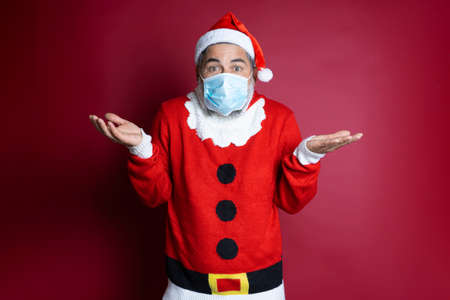 Portrait of man with mask cap and christmas sweater spreading hands with surprise over red backgroundの写真素材
