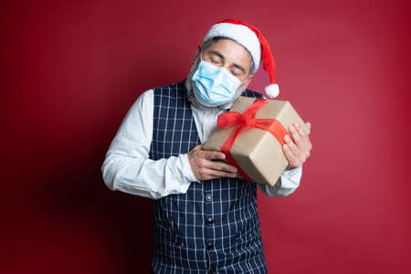 Hipster man with surgical mask and christmas hat emotionally holds a gift box on red background