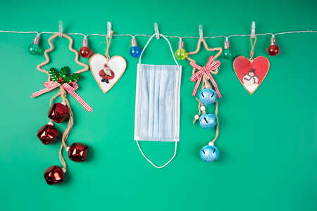 Christmas decorations and a medical mask hang from a garland of colored lights against a green...