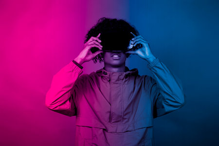 Afro boy with VR glasses. A young afro immersed in virtual reality makes movements with his hands. Blue and red light falls on a boy.の写真素材