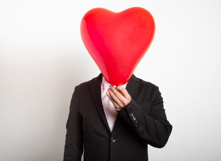 Man in suit hiding behind a red heart shaped balloon. Man holding symbol of love, family, hope.の写真素材