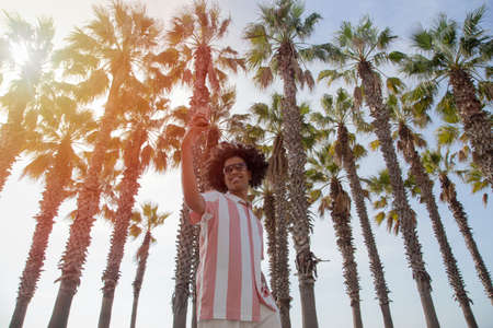 Latino in his 20s taking a selfie among the palm trees on the beach. Dark-skinned man taking a...