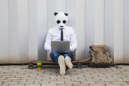 Businessman wearing panda mask and laptop while sitting on skateboard against gray wall.の写真素材