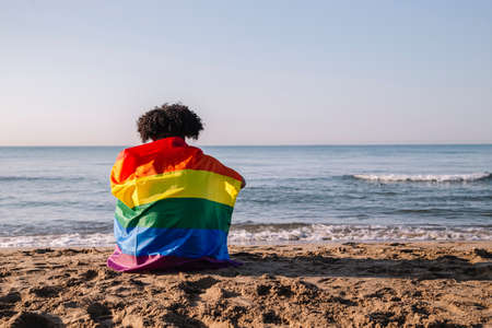 Young latino man with afro hair sitting on the shore of the beach with a lgtbi flagの写真素材