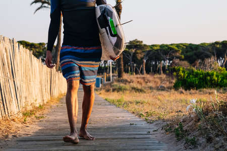 Unrecognizable surfer walks down the wooden walkway to the beach with a surfboard at sunrise.の写真素材