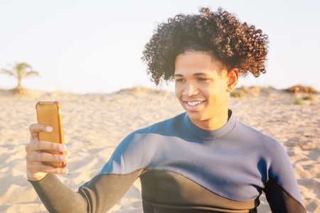 Young African American man sitting on the sand takes a picture with a smartphoneの写真素材