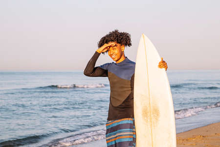 Happy young surfer with surfboards looking to the horizon on the beach at sunrise.の写真素材