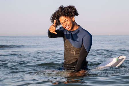 Surfer with afro hair sitting on his surfboard in the sea water sipping and doing the surfer salute...