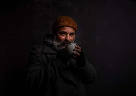 Bearded beggar man smiling joyfully with cup of hot tea isolated on black background