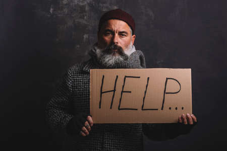 Sad exhausted homeless man in red cap and street clothes holding a cardboard sign with the word help posing in the studio on a dark background.の写真素材