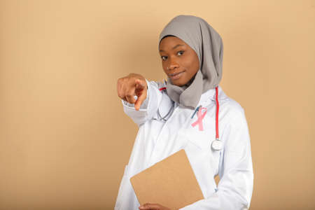 Photo of Muslim female doctor pointing with index finger towards camera wrapped in gray veil and folder with sad face.の写真素材