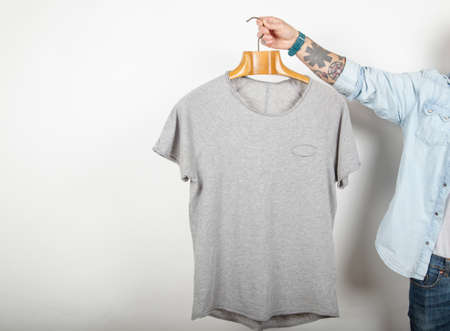 Tattooed male hand holds a hanger with premium quality fine cotton gray t-shirt on a white background. Mockup.の写真素材