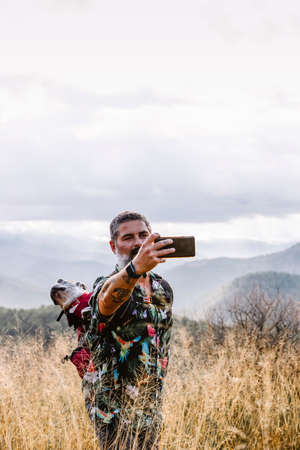 man with dog in backpack taking photos on the mountain with cloudy sky