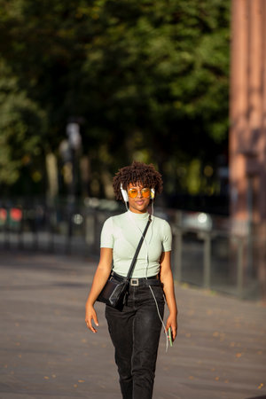 Stylish dark-skinned curly young brunette in yellow sunglasses enjoys music in headphones while walking on the streetの写真素材