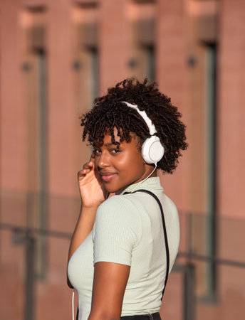 Attractive Latin American teenage girl with curly hair listening to music with white headphones on the street. From the back looking at the cameraの写真素材