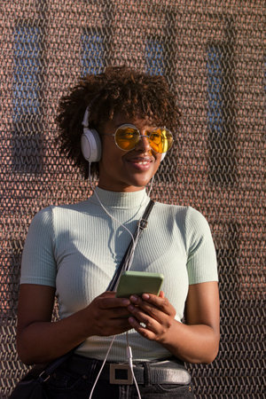 Cool young latin american woman with afro hair and glasses smiling listening to music on smartphone with headphonesの写真素材