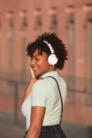 Latin American teenage girl with curly hair listening to music on smartphone wears headphones on the street.の写真素材