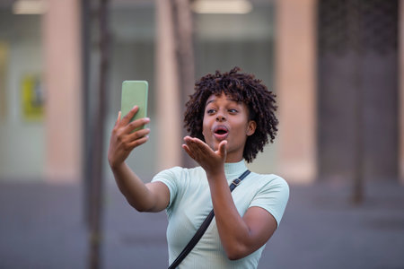 Happy female blogger with afro hair holding phone having video call while walking in the cityの写真素材