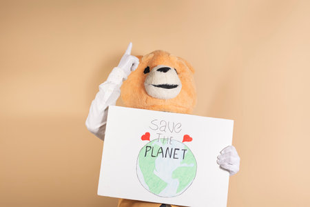 A man in a teddy bear mask on a yellow background points up while holding a cardboard sign that reads, "Save the planet."の写真素材