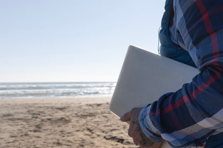 Man's hand holding a laptop on the beach, working outdoors, digital nomad lifestyle concepts