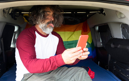 middle aged gay man sitting inside a camping van with lgtbi flag gesturing with a smartphoneの写真素材
