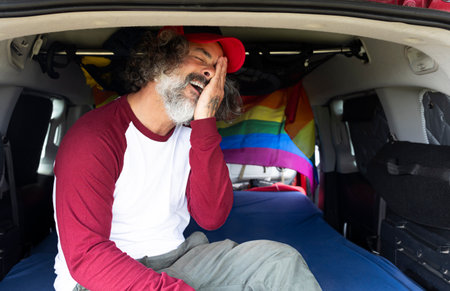Happy gay man in cap sitting inside a camping van with lgtbi flagの写真素材