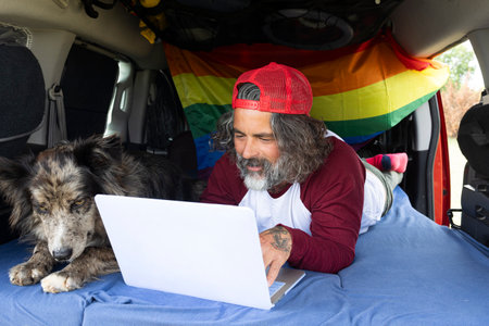 Middle-aged man lying in the camping van with his pet and using a laptop. A traveler with a remote...