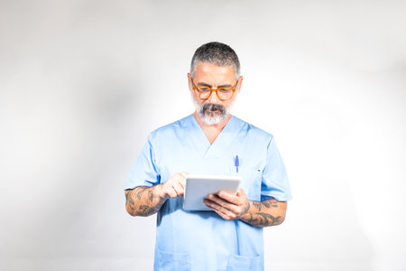 Mature male doctor wearing blue scrubs and glasses, using digital tablet, providing efficient and modern healthcareの写真素材