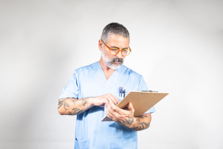 Experienced male doctor in blue scrubs meticulously reviewing medical records on a clipboard, showcasing healthcare expertise and patient careの写真素材