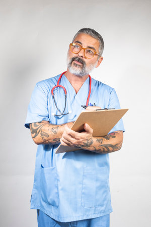 Pensive doctor with tattoos wearing blue scrubs and stethoscope writing notes on clipboard while thinkingの写真素材