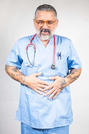 Mature doctor experiencing stomach or abdominal pain, holding his belly with both handsの写真素材