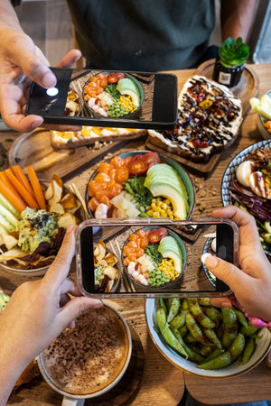 People hands holding smartphones to capture healthy poke bowls and other food on a wooden tableの写真素材