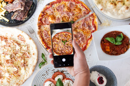 Hand holding smartphone capturing vibrant top down view of a table full of different dishesの写真素材