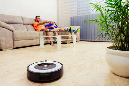 Berlin, Germany, August 29, 2019. irobot roomba cleaning the living room floor while two men are sitting on a sofa. Relaxing momentのeditorial素材