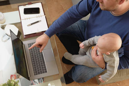 Young father working from home while holding his baby on his lap as they both look at the screen
