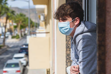 old elderly man in protective face mask against virus looks out through window stay at home for...