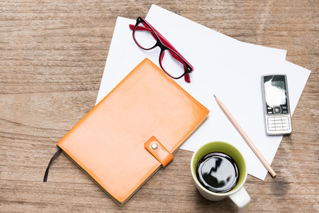 Top view orange diary,blank paper with pencil, a cup of coffee, mobile phone and eye glasses on wood table.の写真素材