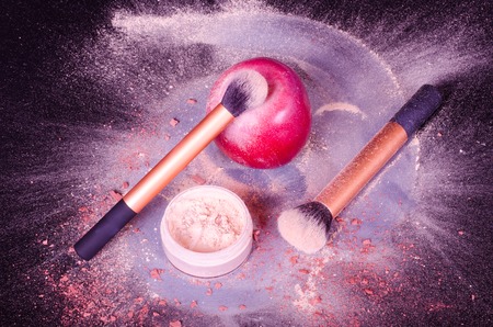 Face powder, two brushes, and red apple on black background